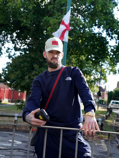 Man standing by a fence holding a phone, with an English flag in the background.
