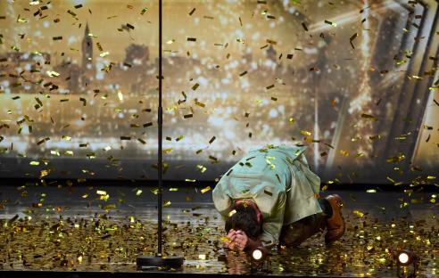 Max Fox kneeling on stage amidst falling gold confetti.