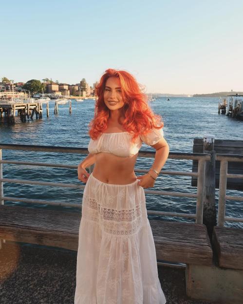 Woman with orange hair wearing a white crop top and skirt by the water.