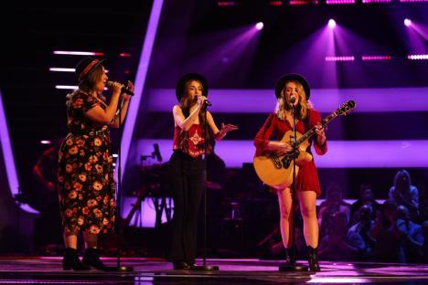 Three women singing on stage, one playing an acoustic guitar.