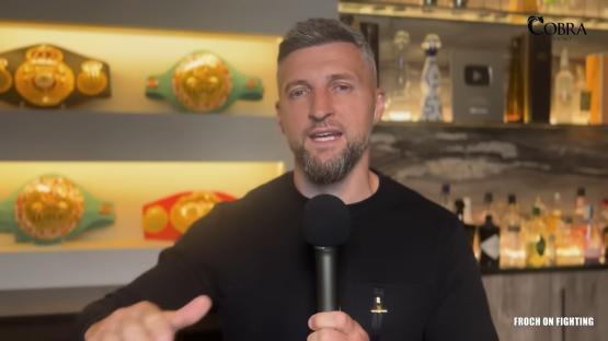 Carl Froch speaking into a microphone. Boxing belts are visible in the background.