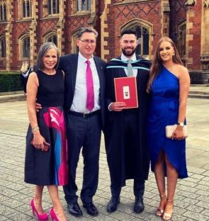 Family photo in front of a university building; a graduate in cap and gown stands with his family.