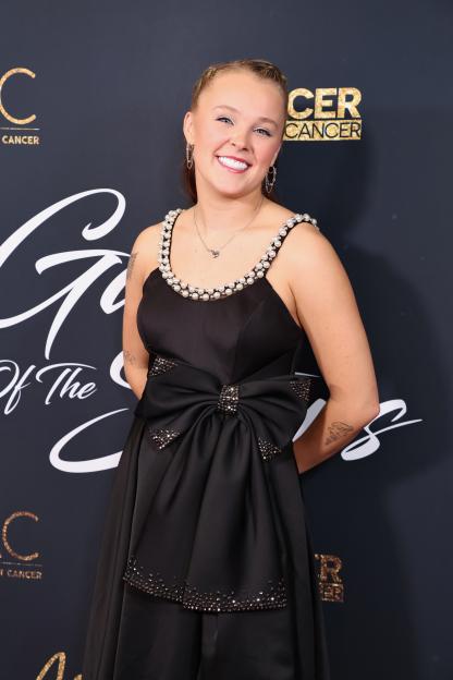 JoJo Siwa smiling in a black dress with pearl-studded straps and a large bow detail.