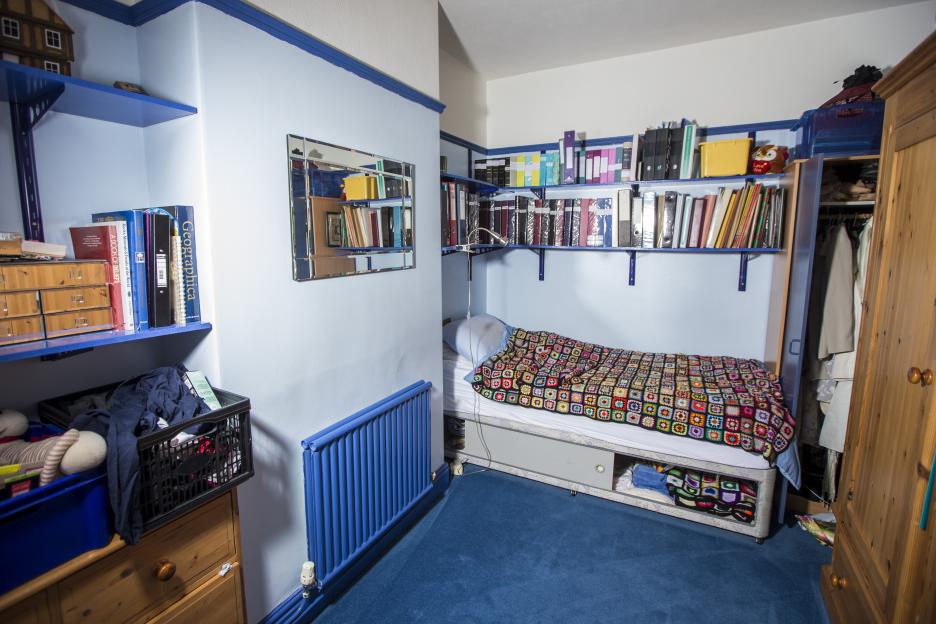 Andrew Gosden's bedroom, with a bed covered by a colorful granny square blanket, shelves of books and binders, and a wooden wardrobe.