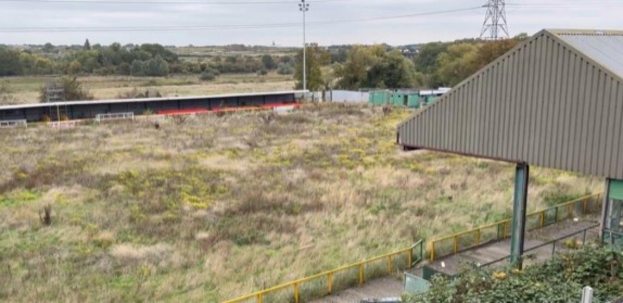 An overgrown football pitch with a small covered stand to the right and a long, low stand in the background.