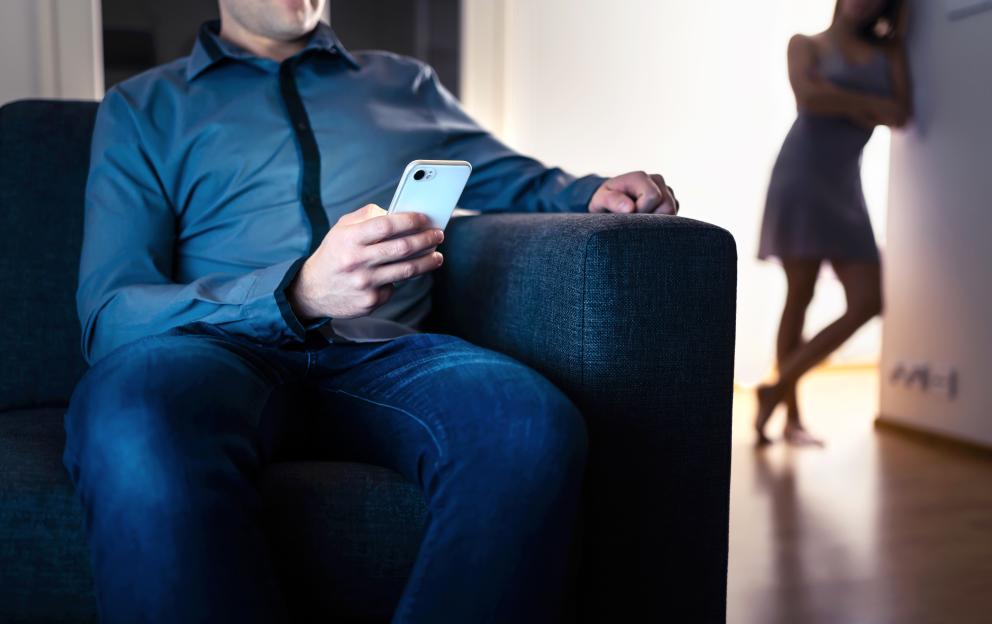 A man sits on a couch using his phone while a woman stands in the background.