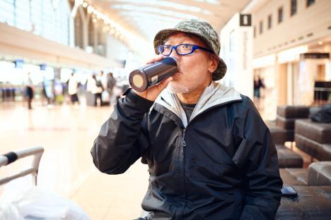 He is wearing a thin black jumper, a hat, and drinking water from a water bottle while waiting to board.
