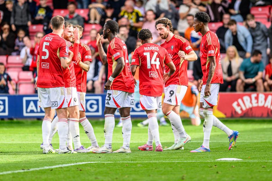 Salford City FC players, including Cole Stockton (not visible but inferred from meta-data), celebrating a goal with high-fives and huddles during a soccer match.