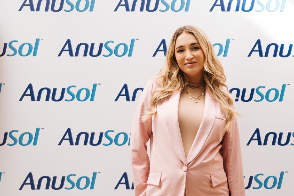 A woman in a pink blazer standing in front of a white wall with the Anusol logo repeated.