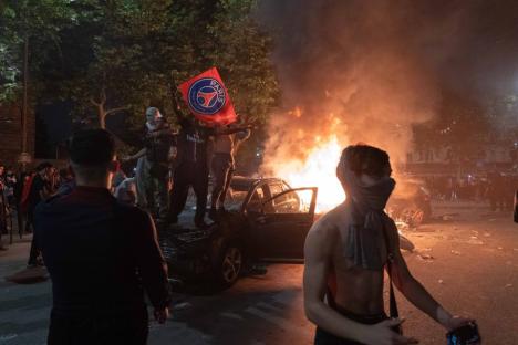 A burning car during a riot; people are standing on the car, one holding a Paris Saint-Germain flag.