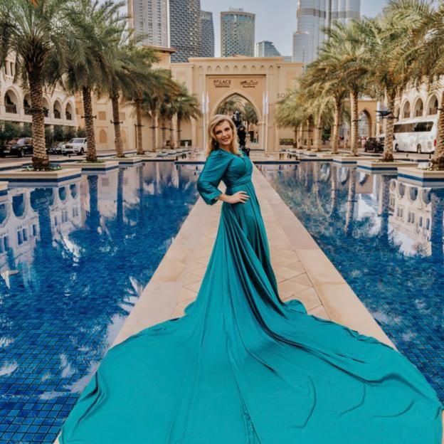 Anastasiia Chikina, 32, a Russian mother, standing next to a large pool with palm trees, buildings, and an ornate archway in the background.