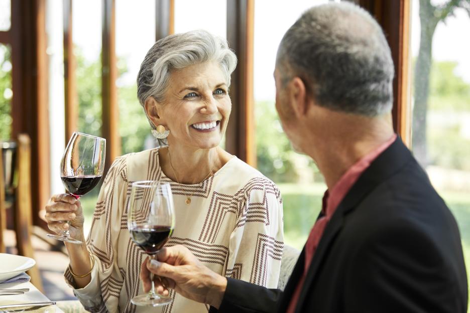 A smiling senior woman with red wine looks at her male partner at a restaurant table.