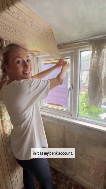 A smiling woman in a white t-shirt and black leggings peels back wallpaper from a ceiling, revealing a window behind her with a "For Sale by Auction" sign.