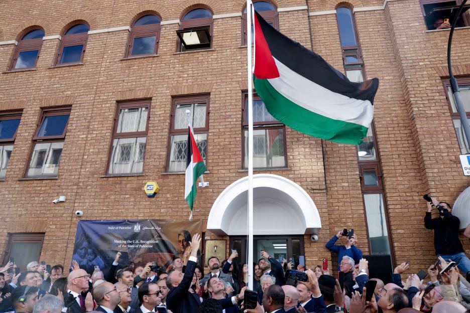 The Palestinian flag is raised at the Palestinian mission in London, watched by a crowd of people.
