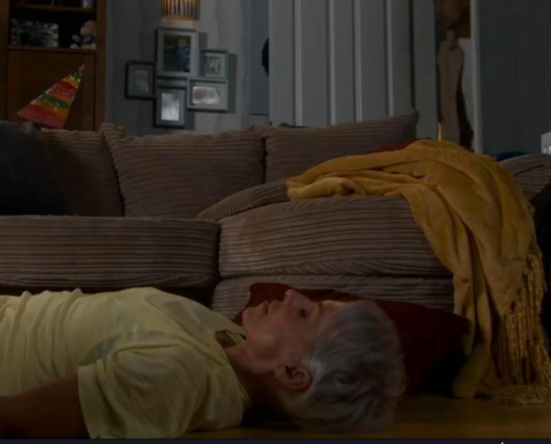 Screenshot of an elderly woman lying on the floor near a couch.