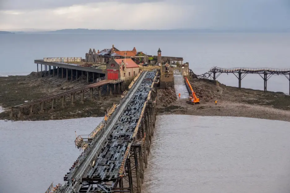 Abandoned pier with buildings on an island, with a digger and person on the shore.