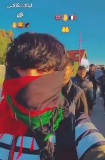 A person wearing a red, green, and black scarf stands with a group of people near Calais and Dover.