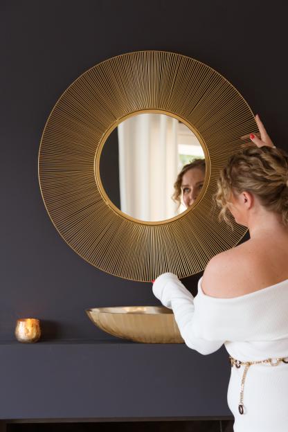 A woman in a white off-shoulder dress is reflected in a gold round mirror with sunburst rays.