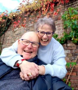 Smiling couple embracing in front of a brick wall covered in autumn leaves.