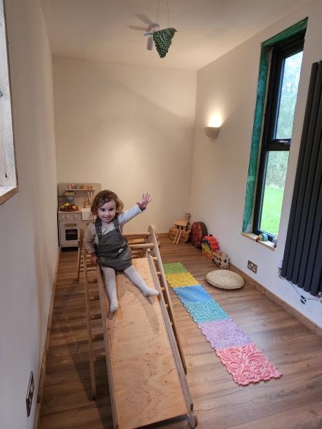 Toddler Naya sits on a wooden slide in a tiny home with a toy kitchen, play mat, and outdoor view.