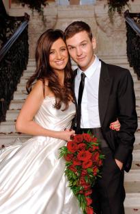 A bride and groom pose on a staircase, holding a bouquet of red roses.