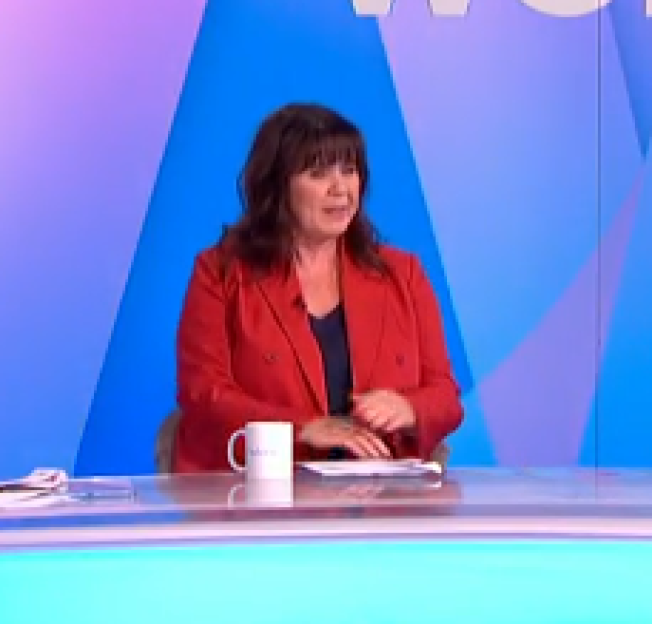 Screenshot of a woman in a red jacket sitting at a desk and speaking.