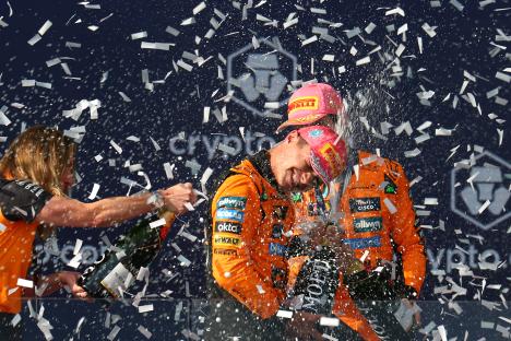 Lando Norris and Oscar Piastri celebrating on a podium with champagne and confetti.