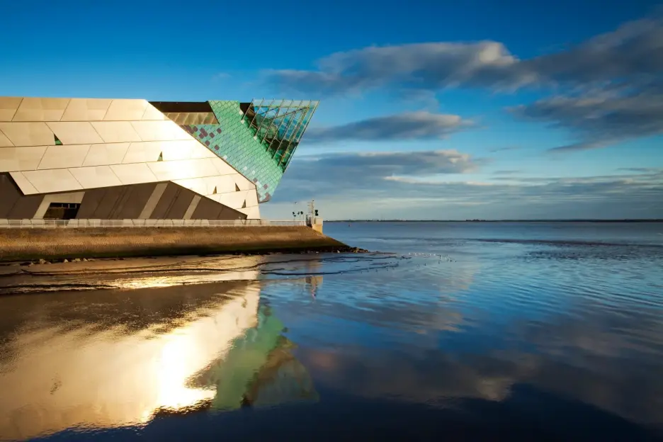 Modern building with a metallic facade and green glass extension reflected in a calm body of water under a blue sky with clouds.