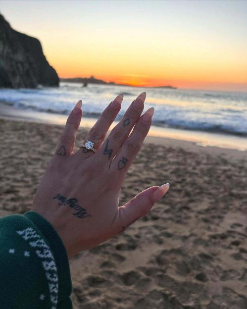 A hand wearing a ring with a large diamond and a band with smaller diamonds, against a beach sunset background.