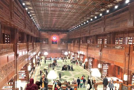 Interior view of a large, ornate library with multiple levels of bookshelves and people browsing.