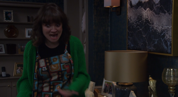 Screenshot of a woman wearing a green cardigan and patterned apron, looking shocked.