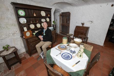 A man sits in a rock-hewn house, surrounded by period furniture and tableware.