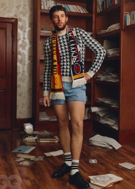Josh O'Connor wearing a patterned sweater, boxer shorts, black shoes, and two scarves, standing in a messy room with bookshelves.
