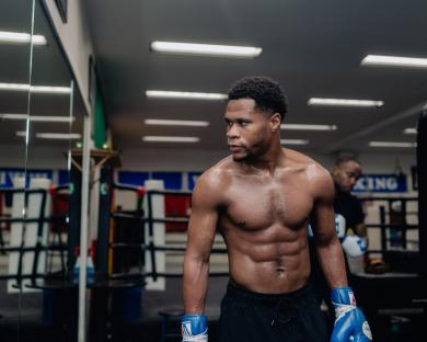 Devin Haney in a boxing gym, shirtless, wearing blue boxing gloves.