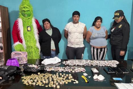 A Grinch-costumed police officer stands with three drug trafficking suspects and a police officer after a drug bust. Confiscated drugs, money, and phones are on the table.