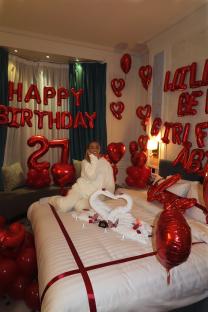 Woman on a bed surrounded by red heart balloons and "Happy Birthday" decorations.