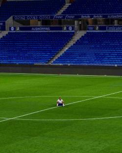 A person sits alone on a soccer field in front of empty stadium seats.