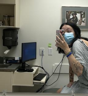 A young woman in a hospital gown, wearing a face mask, holds up her phone to take a selfie in a clinic waiting room.