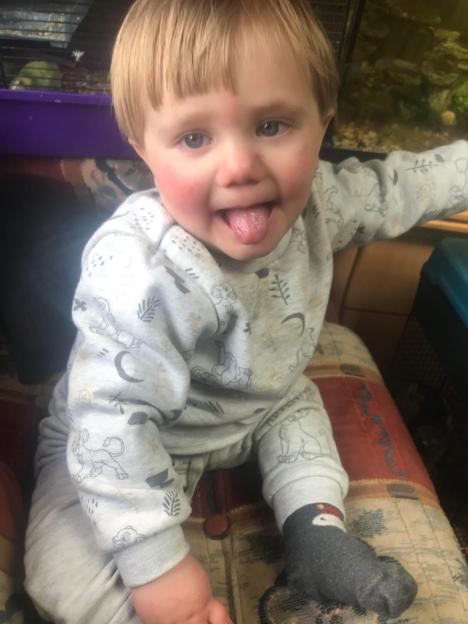 A fair-haired toddler, Cody Davies-Owen, wearing light-colored pajamas and dark socks, sticking out his tongue and smiling.