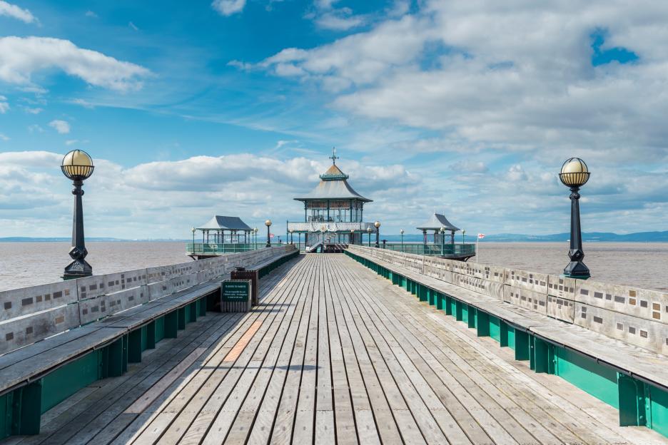 Clevedon Pier, Somerset, England, featuring benches, light posts, and pavilions on a wooden deck extending into the water under a partly cloudy sky.