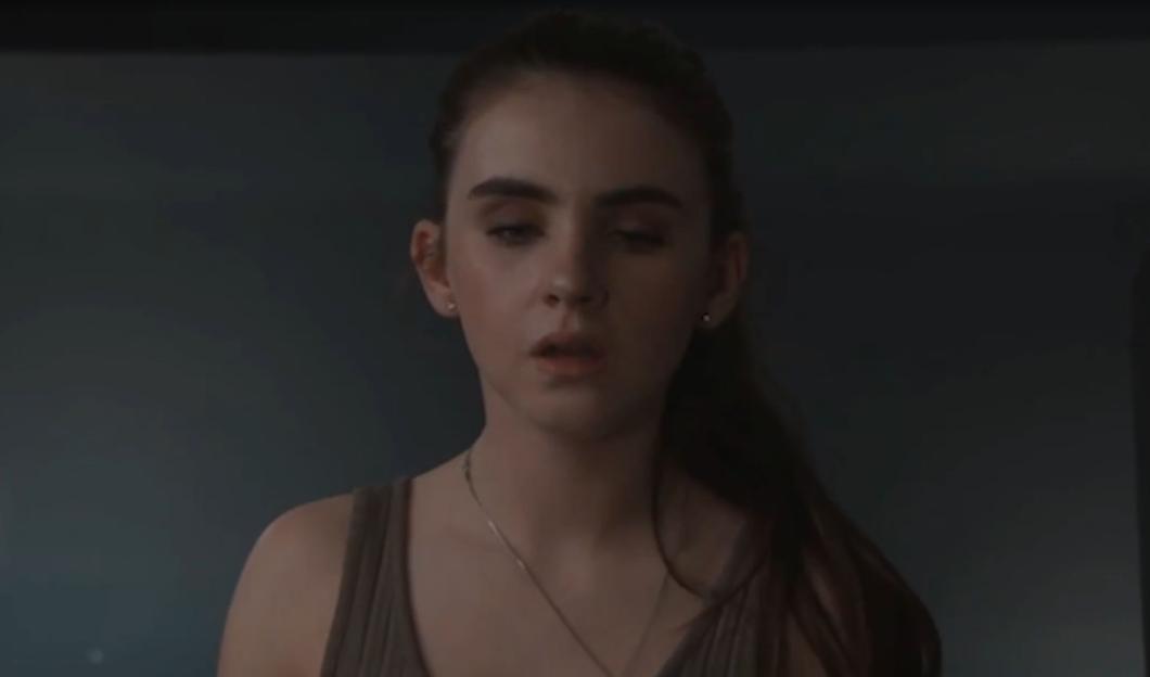 A person with brown hair, in a gray tank top and necklace, looks downward and to the right, with a slightly open mouth.