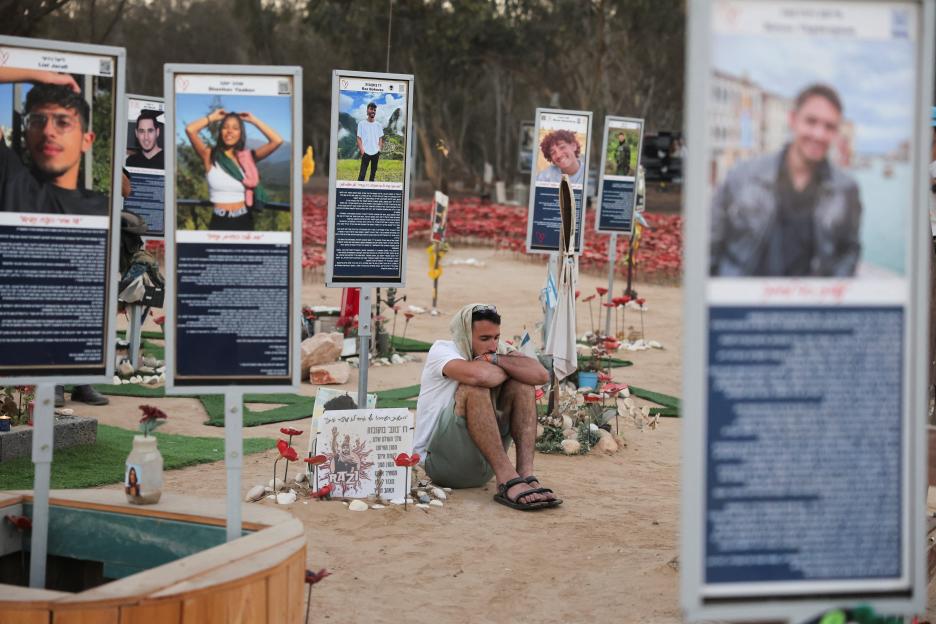 A man sits amidst memorials to Nova festival victims, with portrait boards and flowers surrounding him.