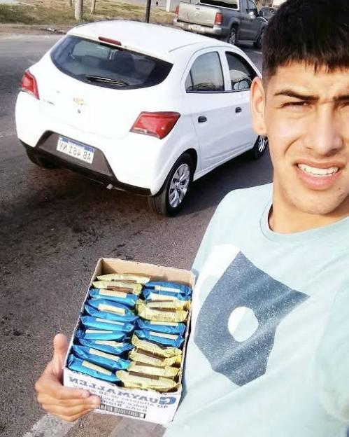 Lautaro Rivero holding a box of packaged foods, with a white car and a truck behind him.