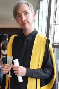 A man in a graduation gown holds a champagne flute and a certificate.