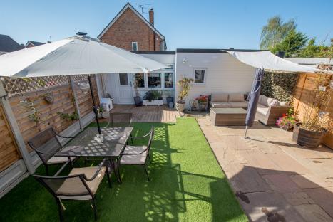 Small sunny backyard patio with furniture, artificial grass, and a parasol.