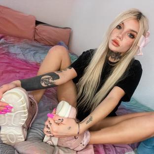 Woman with blonde hair and tattoos sits on a bed and holds up her shoes.