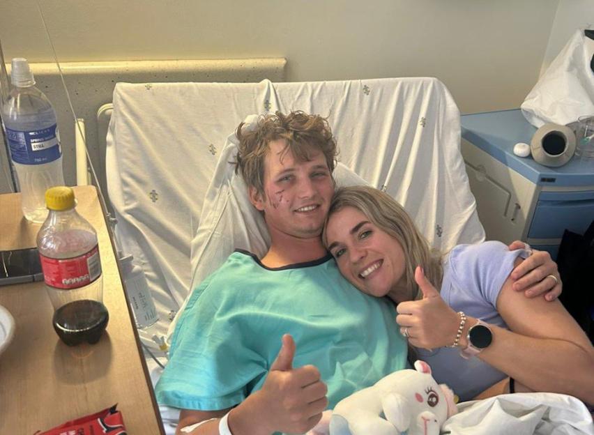 A smiling man in a hospital bed with facial injuries gives a thumbs up with his girlfriend.