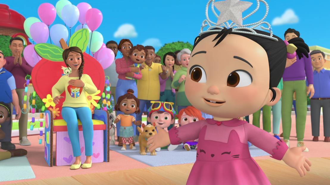 A little girl wearing a tiara stands in front of a crowd of people.
