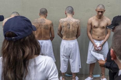 Prisoners with extensive tattoos on their backs, shackled and in white shorts, stand during a prison tour.