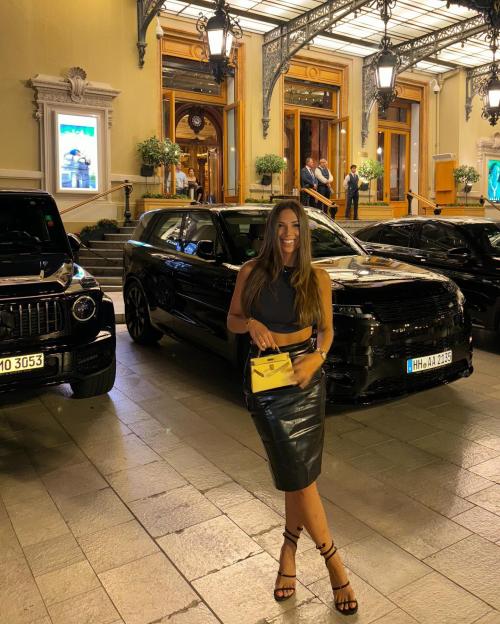 A smiling woman in a black crop top and skirt stands in front of a black Range Rover.